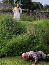 Woman in white dress standing on grassy hill while man in plaid shirt and cap lies on the ground in front, outdoors with stone wall and house in the background.