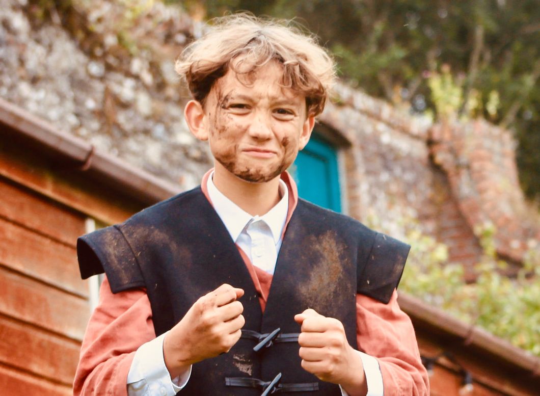 Two people dressed in old-fashioned, rustic clothing, with the child having a dirt-smudged face, standing in front of a red wooden building and a brick wall outdoors.
