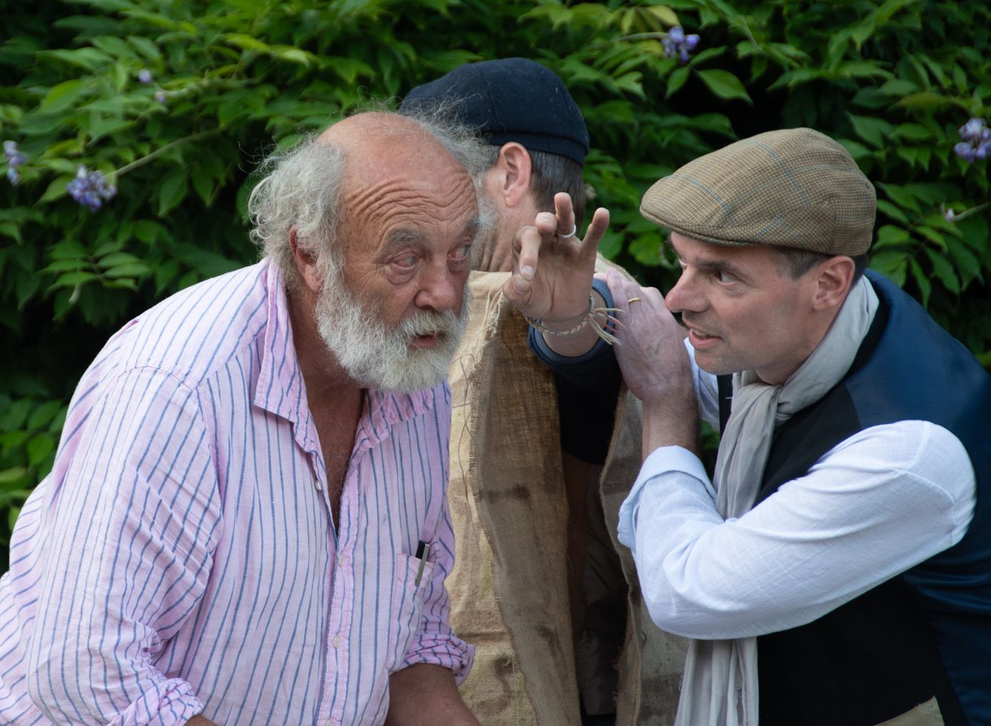 Two men engaged in a lively conversation outdoors with a green leafy background.