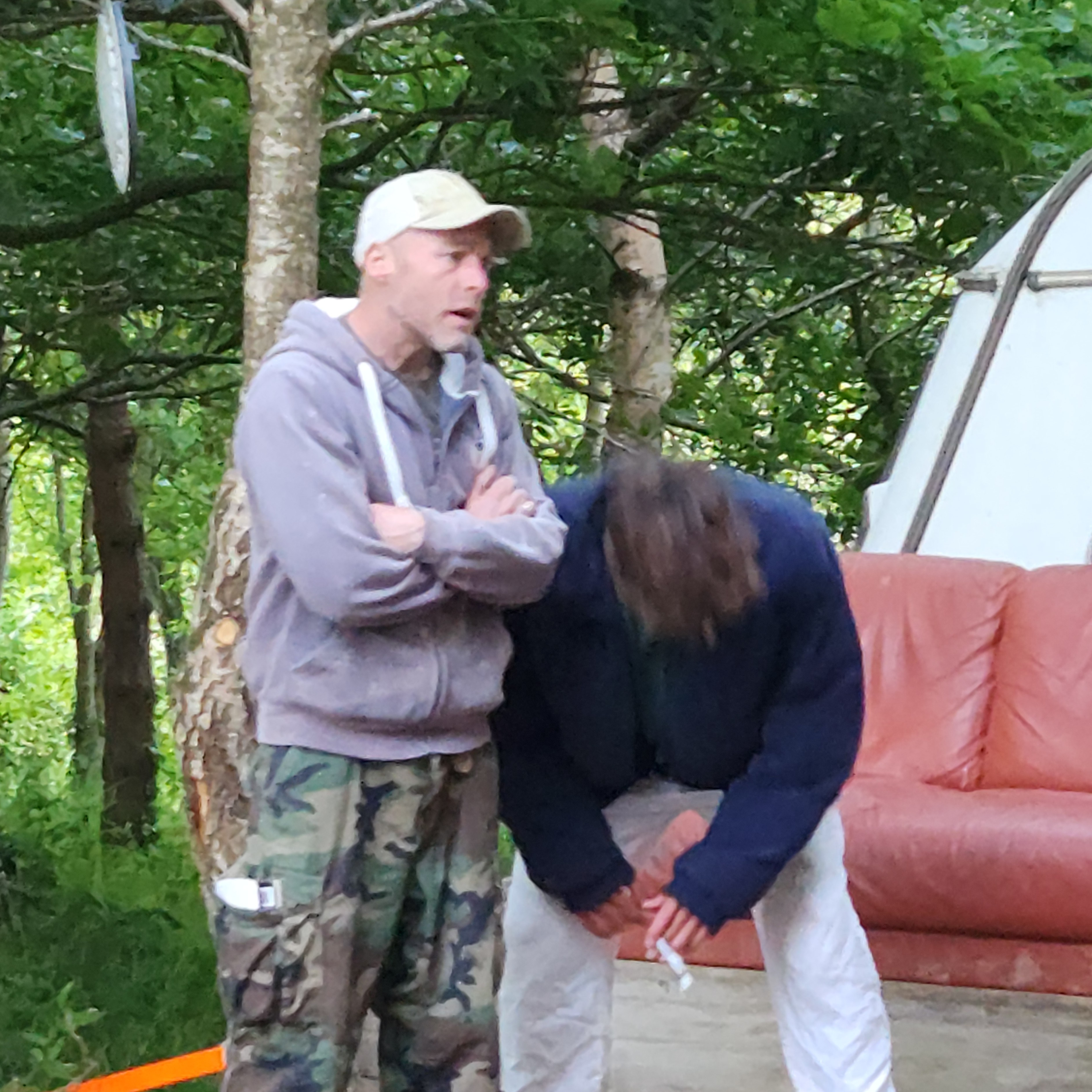 Two people outdoors, one standing with arms crossed and another bending forward, next to a red couch and trees in the background.