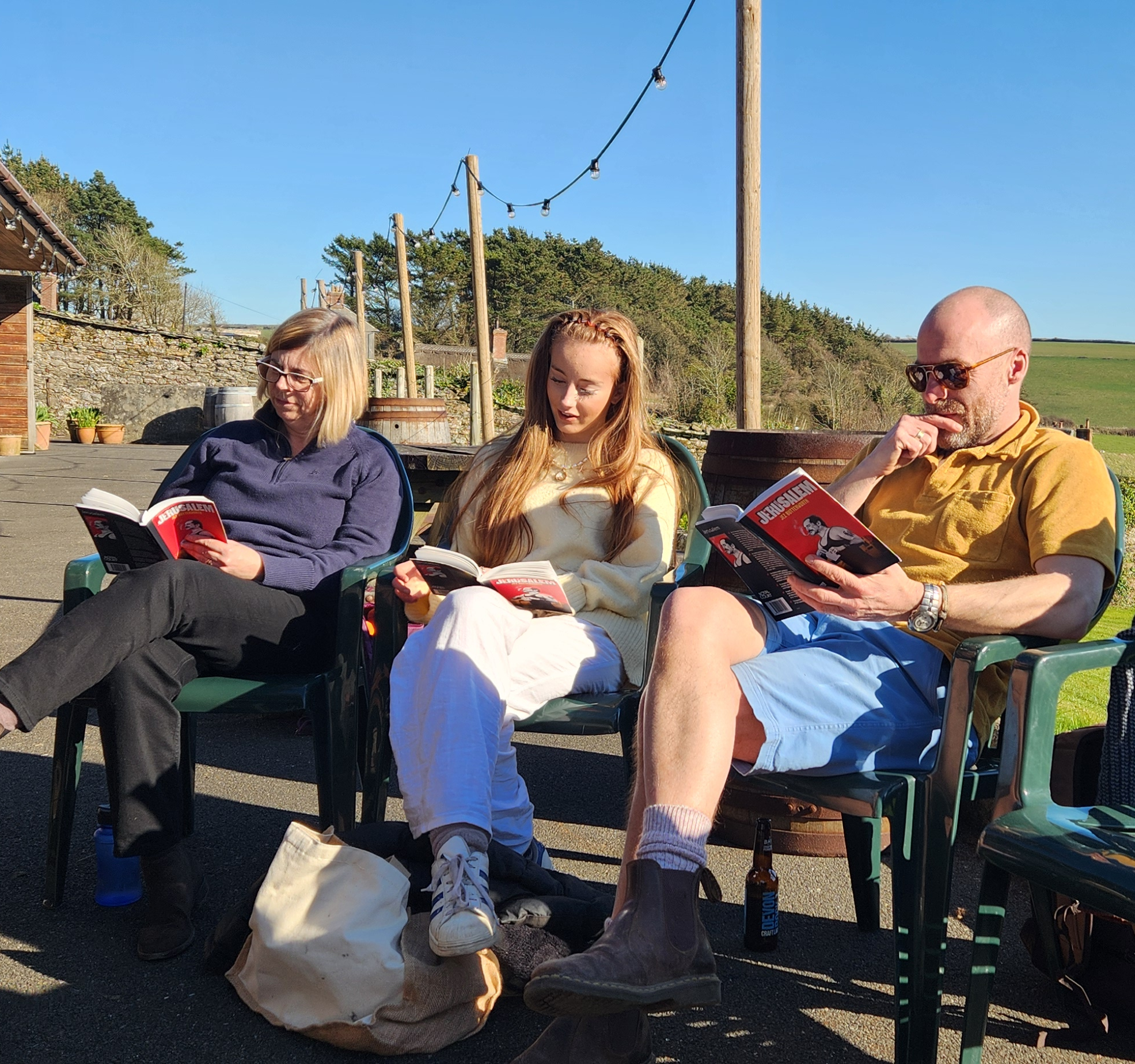 Three people sitting on green chairs outdoors, reading books under a clear blue sky
