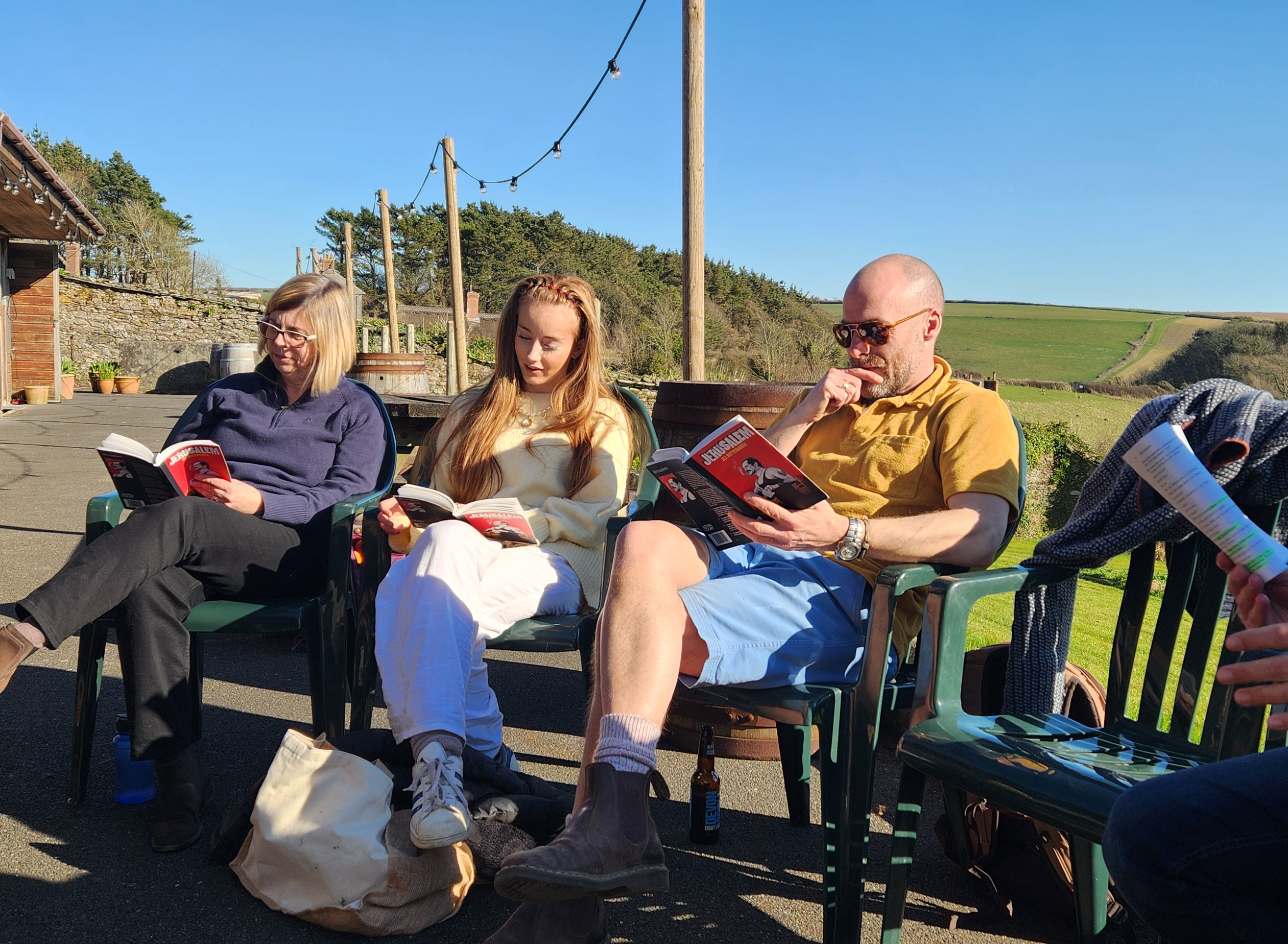 Three people sitting on green chairs outdoors, reading books under a clear blue sky