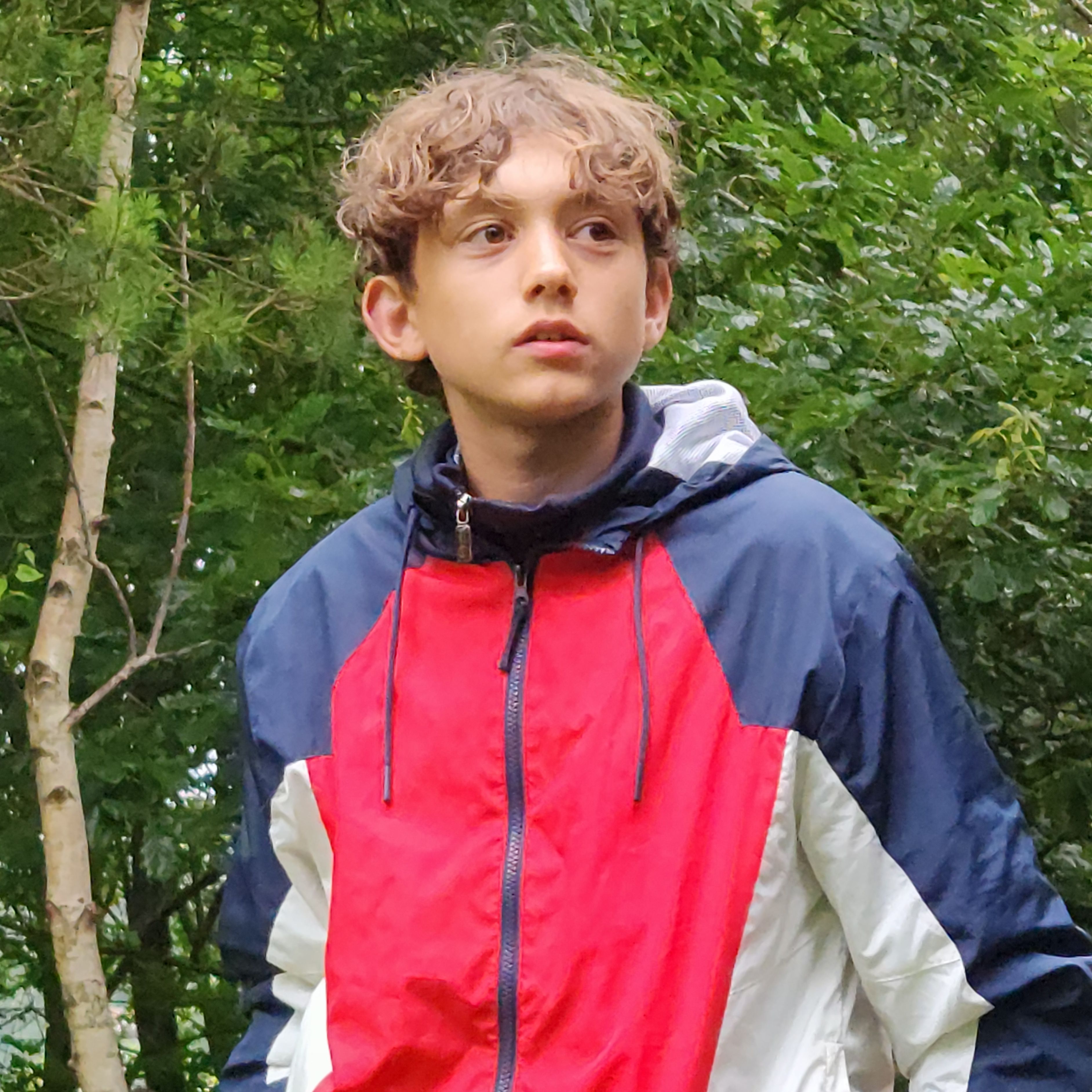 Teenager wearing a red, navy, and white jacket standing outdoors in a wooded area.