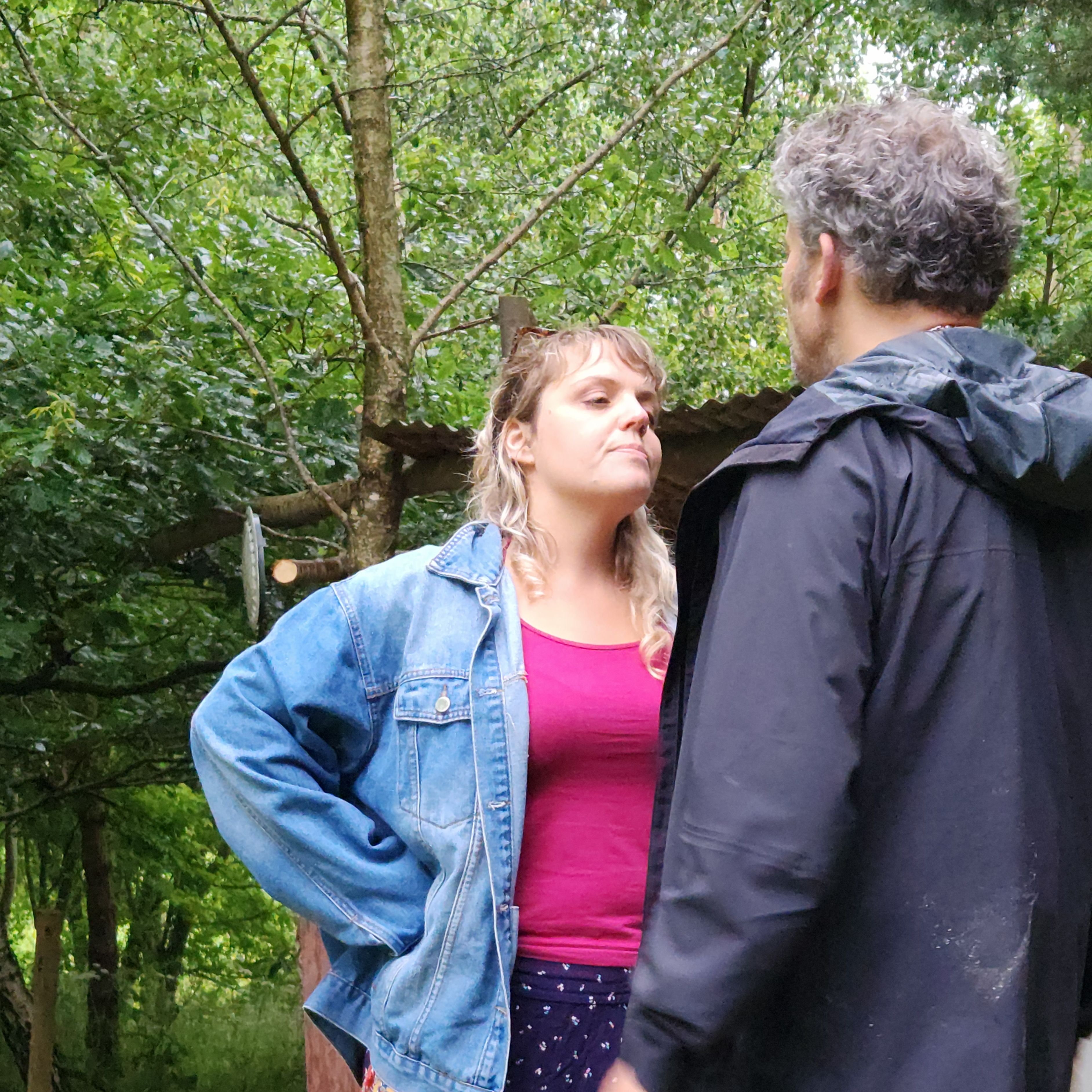 Two people standing and facing each other outdoors, one with hands on hips wearing a denim jacket.
