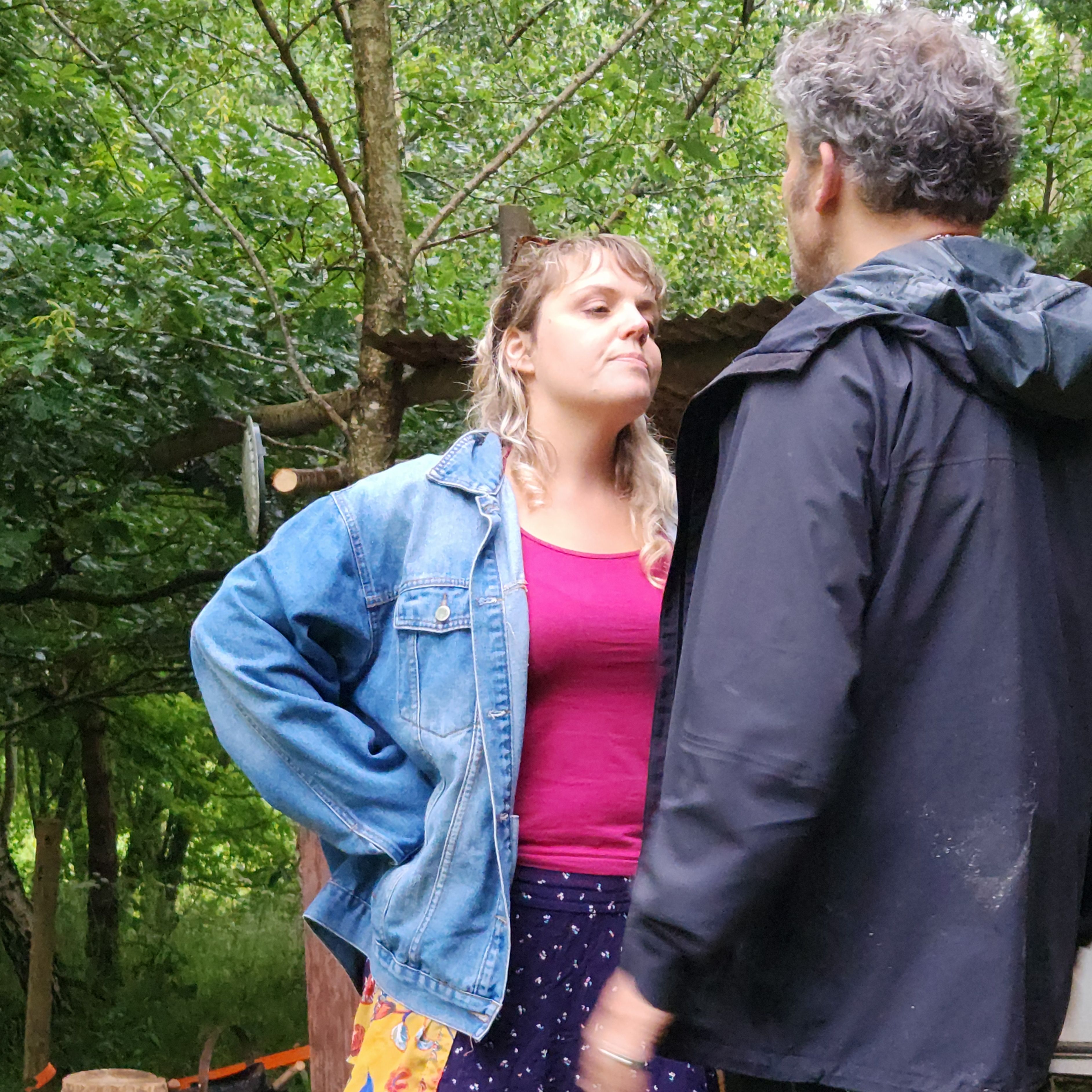 Two people standing and facing each other outdoors, one with hands on hips wearing a denim jacket.