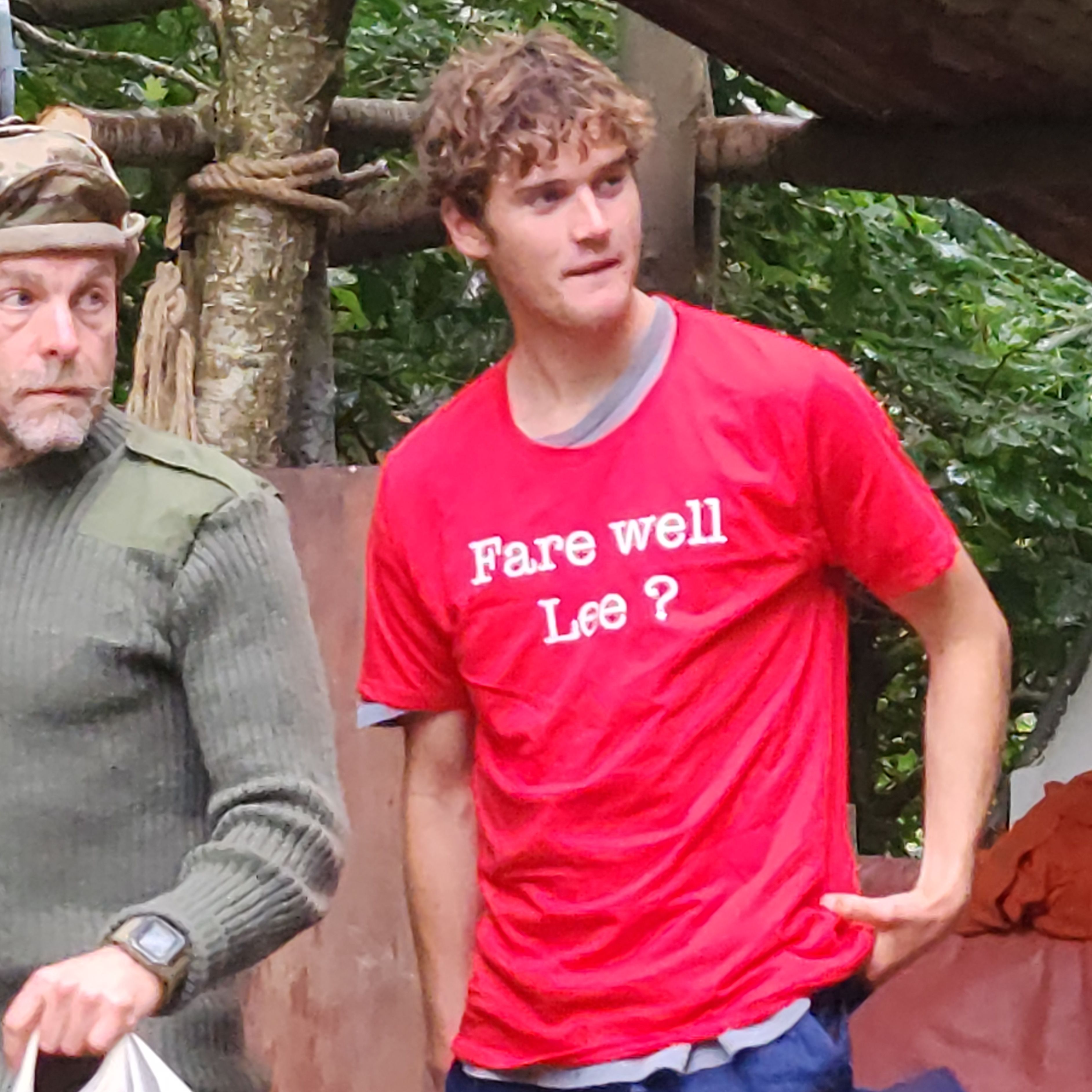 Young man in red t-shirt with 'Fare well Lee?' and blue pants standing outdoors near another person in camouflage.