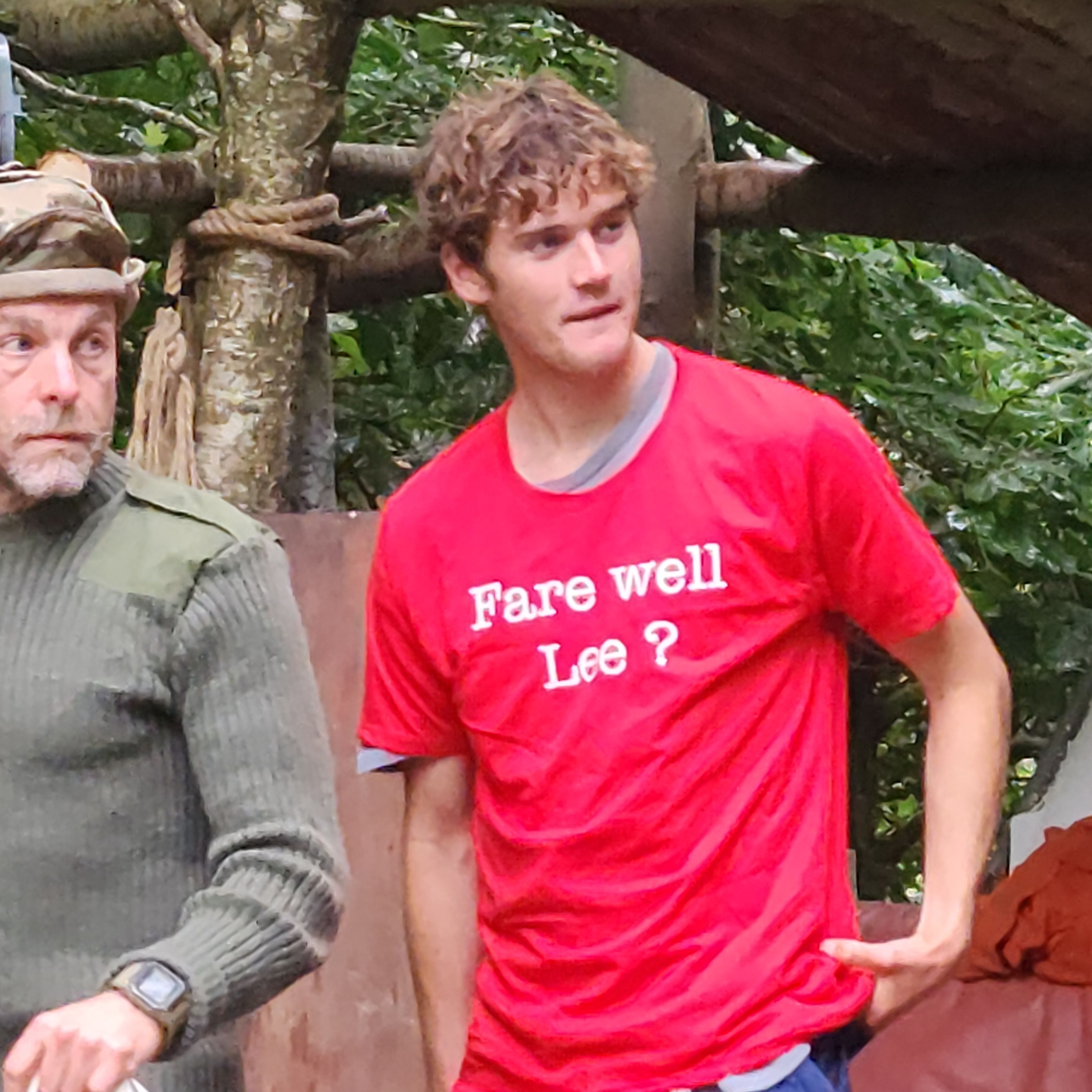 Young man in red t-shirt with 'Fare well Lee?' and blue pants standing outdoors near another person in camouflage.