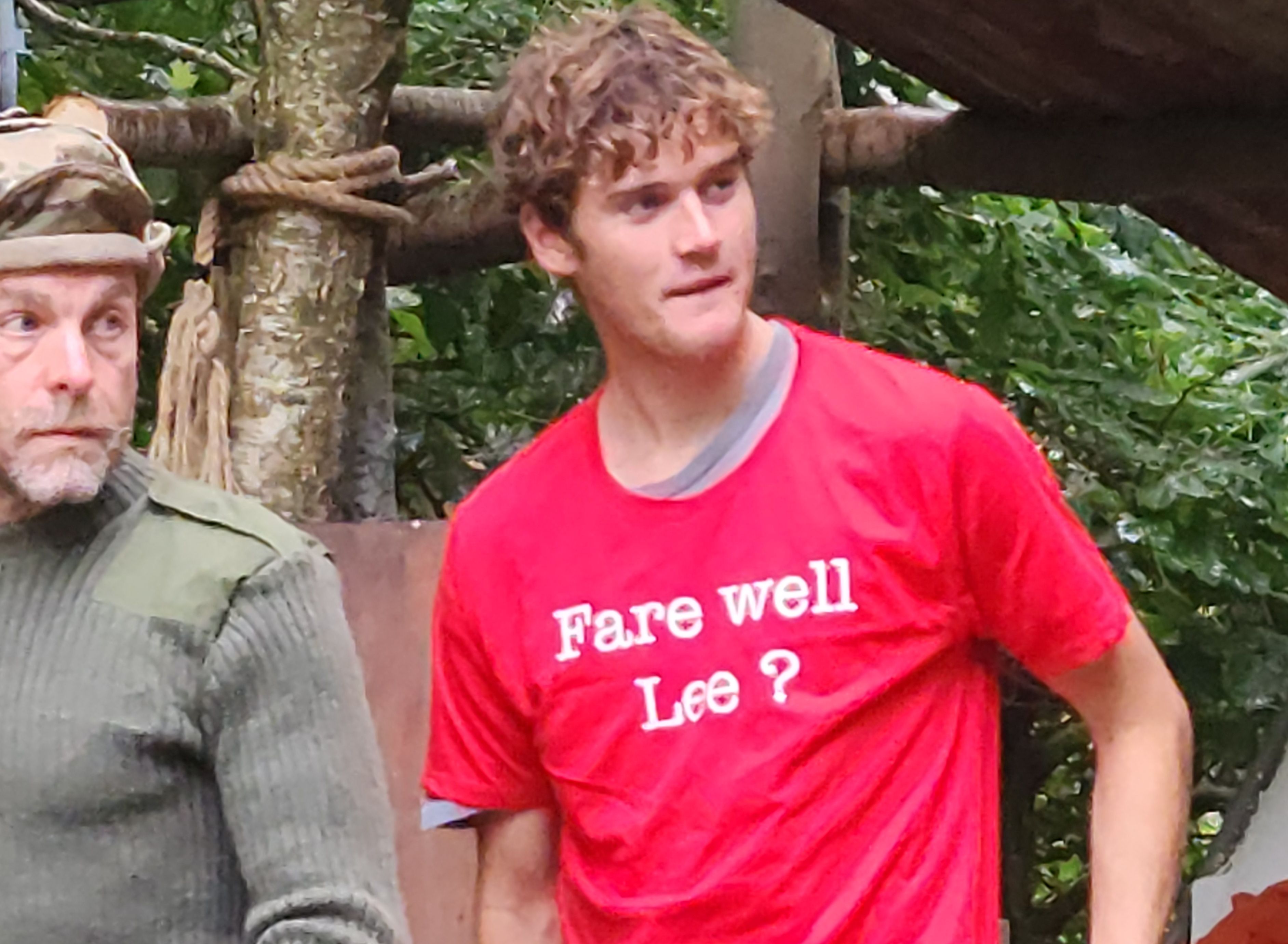 Young man in red t-shirt with 'Fare well Lee?' and blue pants standing outdoors near another person in camouflage.