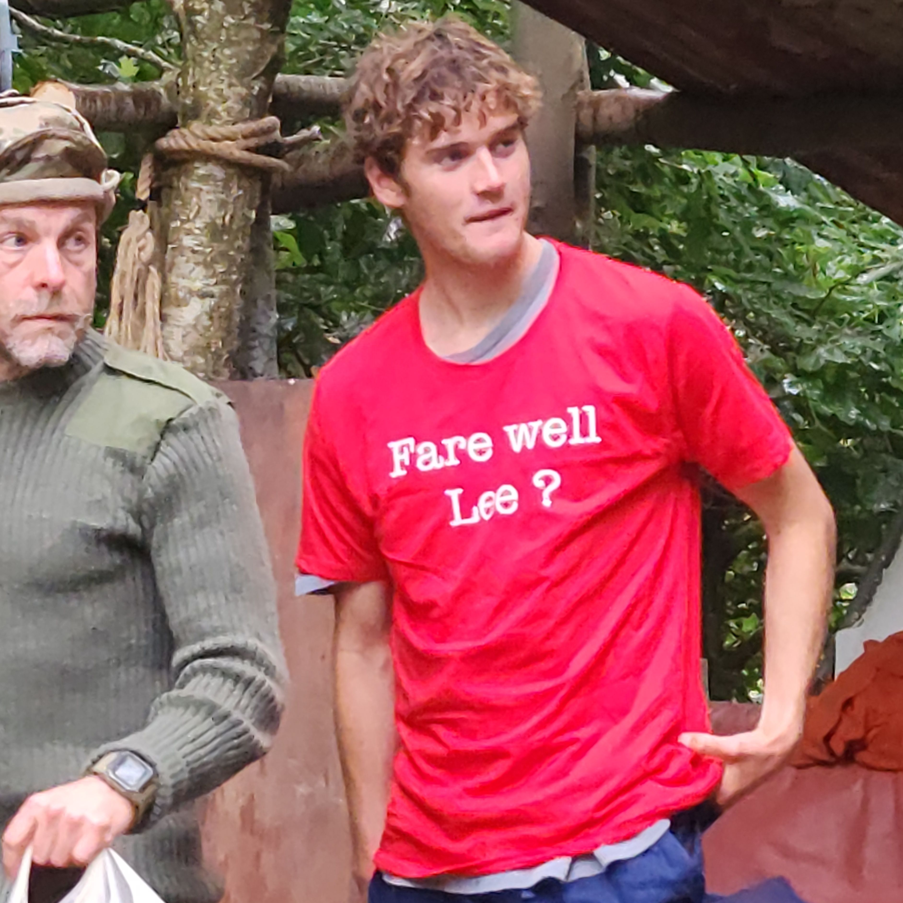 Young man in red t-shirt with 'Fare well Lee?' and blue pants standing outdoors near another person in camouflage.