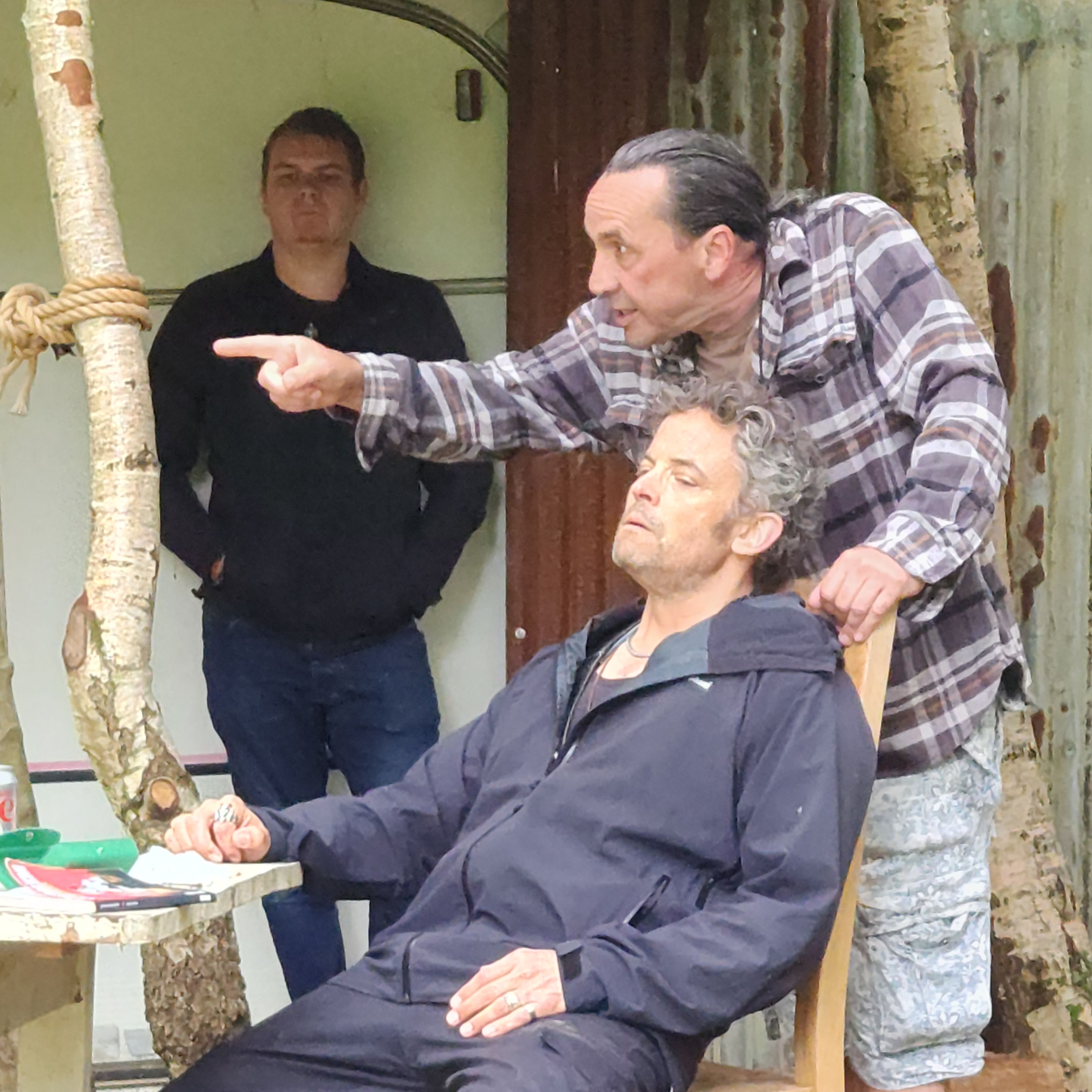 Three men outside near a rustic shelter, one sitting on a chair with eyes closed, another standing beside him pointing, and a third man in the background.