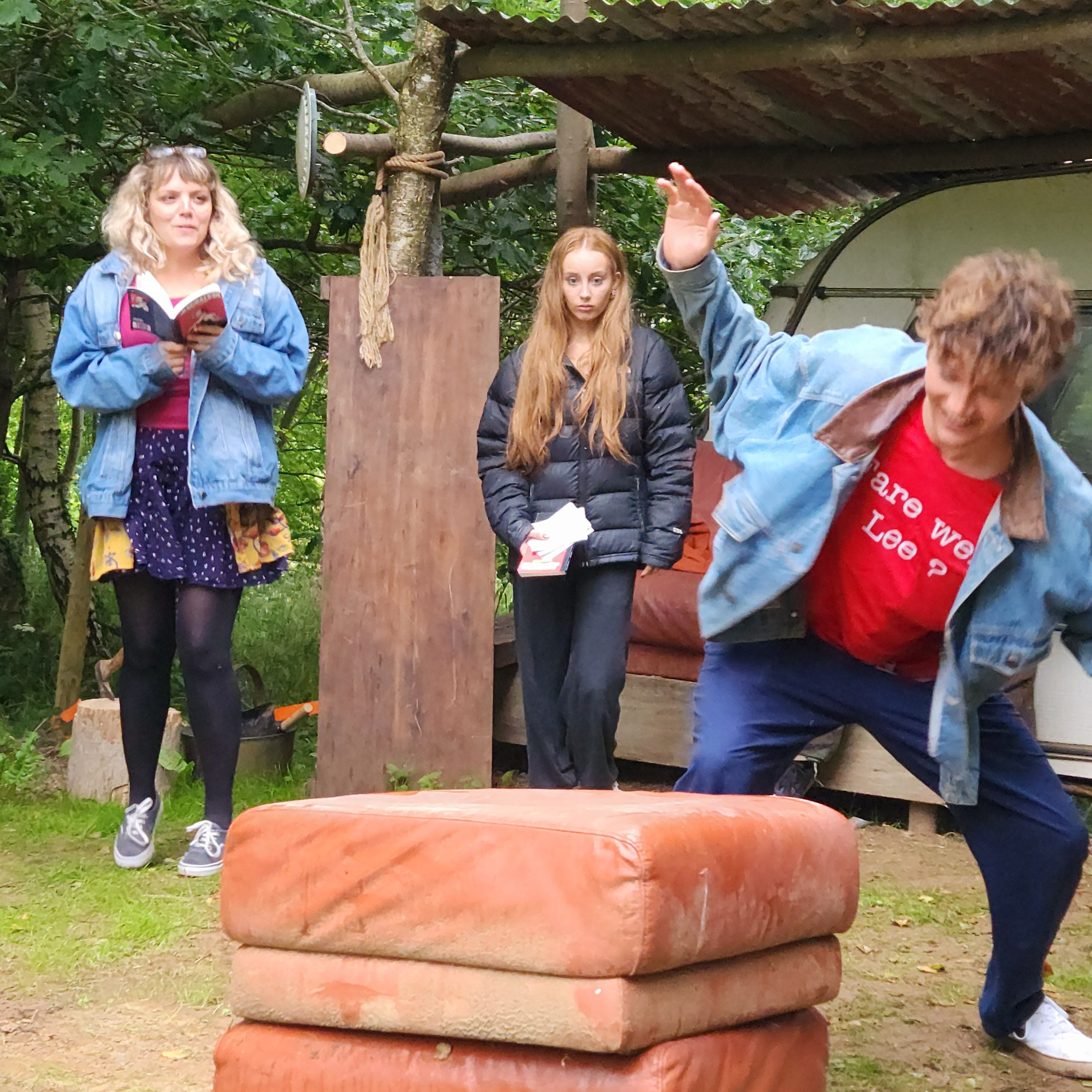Three people outdoors near trees; one is reading, one is standing, and one is leaning forward near a stack of cushions.