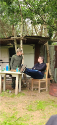 Two men sitting and talking at a table outdoors, surrounded by trees and rustic structures.