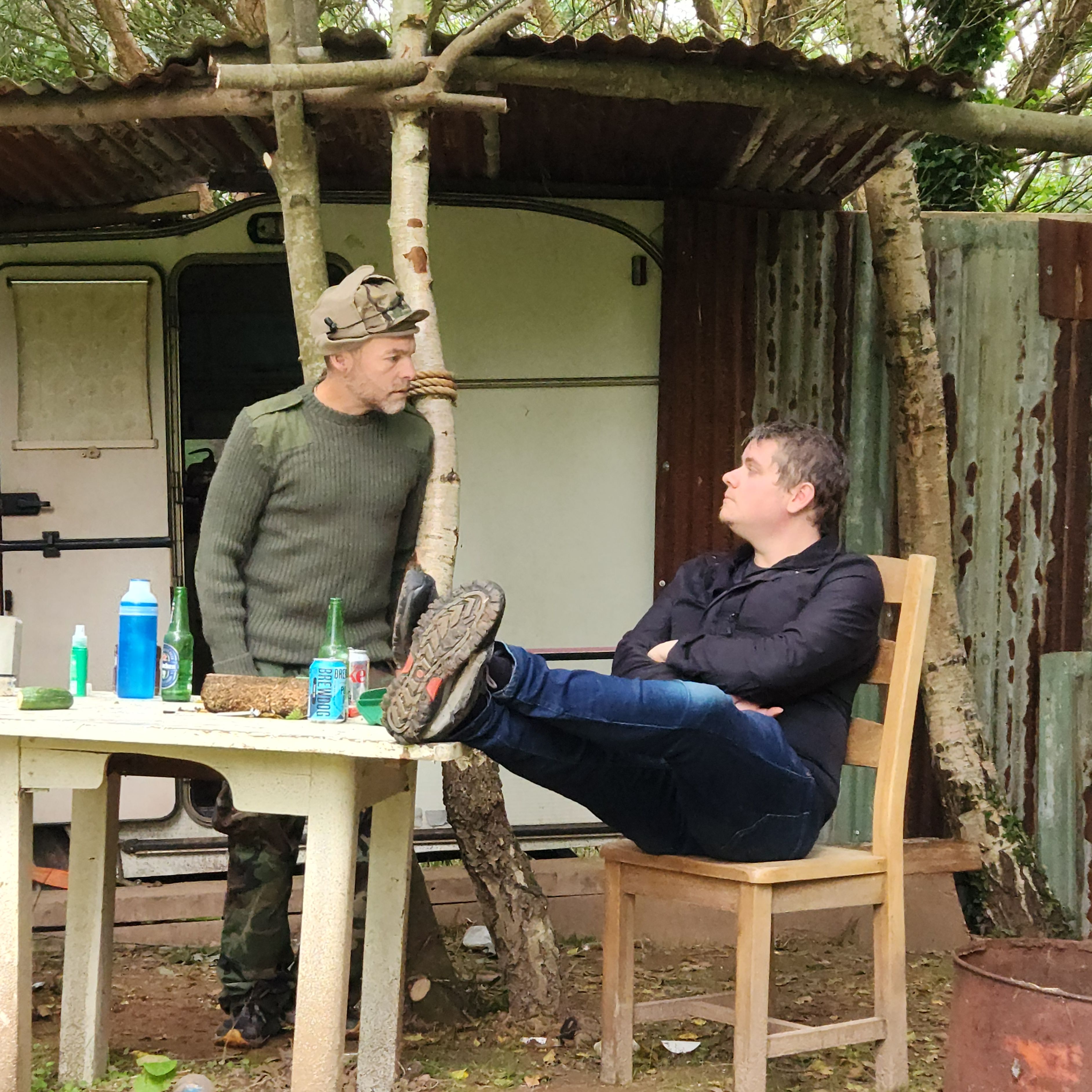 Two men sitting and talking at a table outdoors, surrounded by trees and rustic structures.