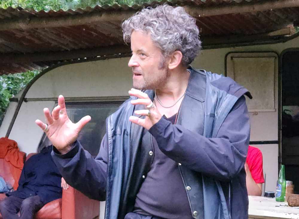 Man gesturing dramatically outdoors near a table and an old couch.
