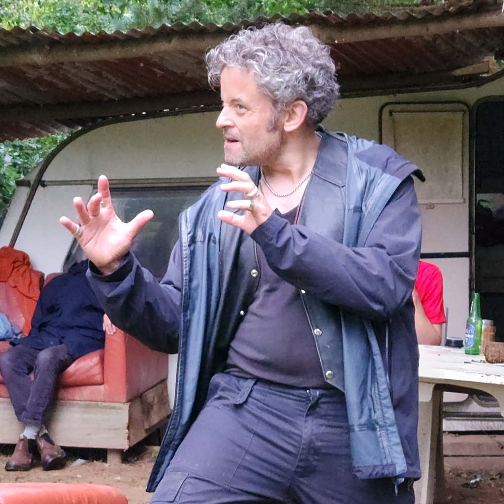 Man gesturing dramatically outdoors near a table and an old couch.