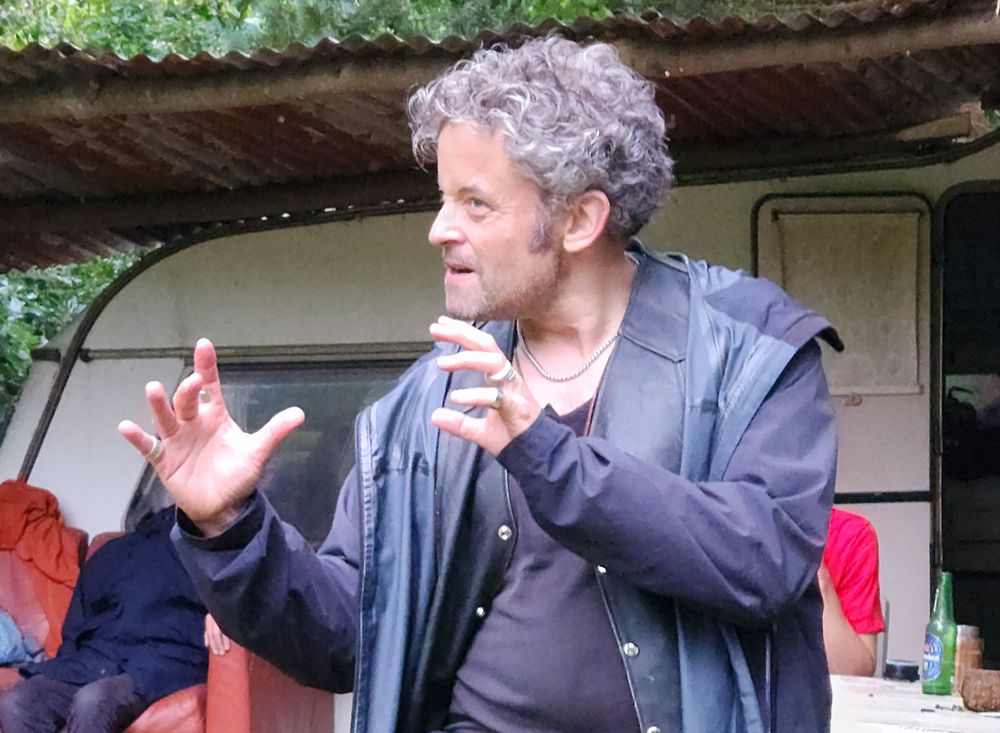 Man gesturing dramatically outdoors near a table and an old couch.
