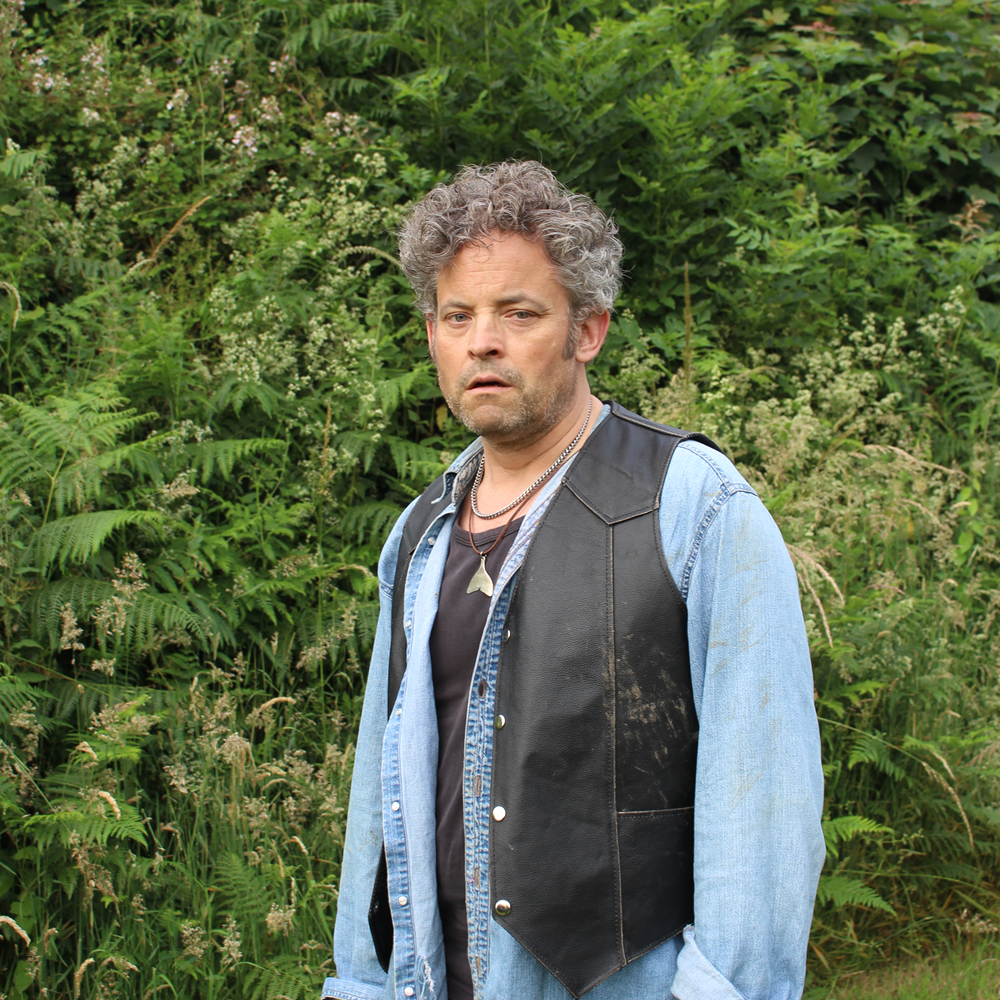 Man with curly hair wearing a denim shirt and black leather vest standing outdoors