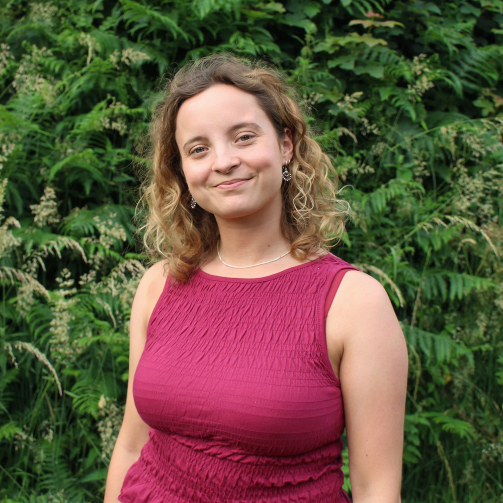 Woman with curly hair wearing a sleeveless maroon dress standing in front of green foliage