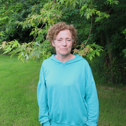 A person with short curly hair standing outdoors in front of leafy green trees, wearing a turquoise hoodie.