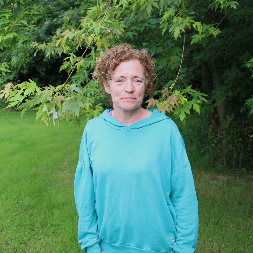 A person with short curly hair standing outdoors in front of leafy green trees, wearing a turquoise hoodie.