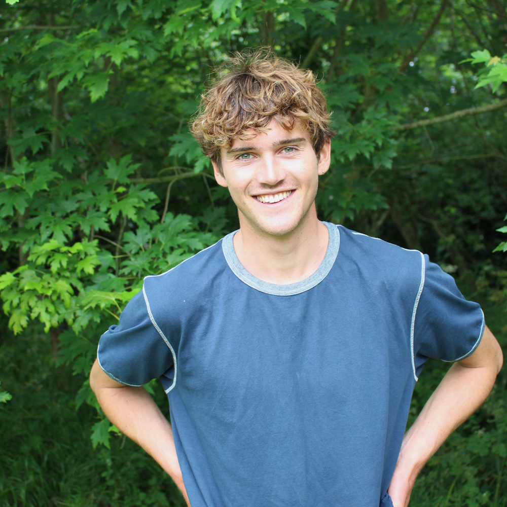 Young man smiling outdoors with hands on hips, standing in front of green leafy trees.