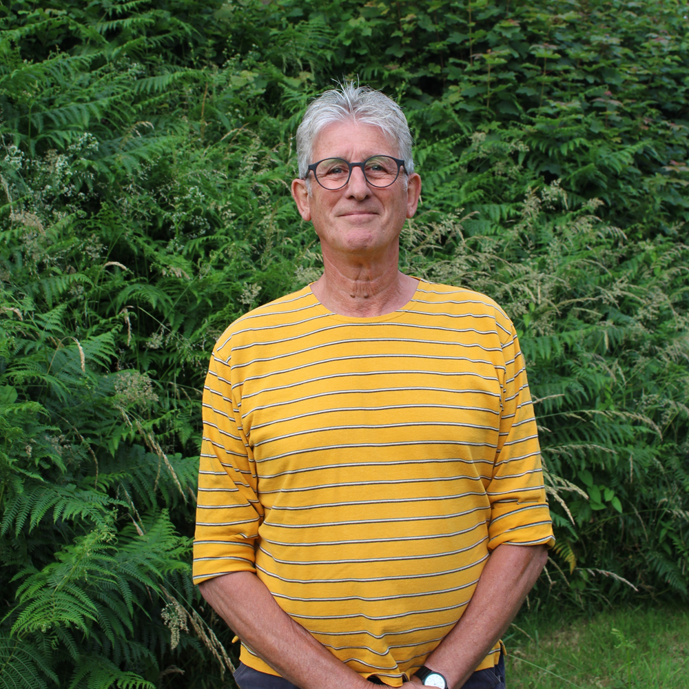Older man with gray hair and glasses wearing a yellow striped shirt standing in front of green foliage.