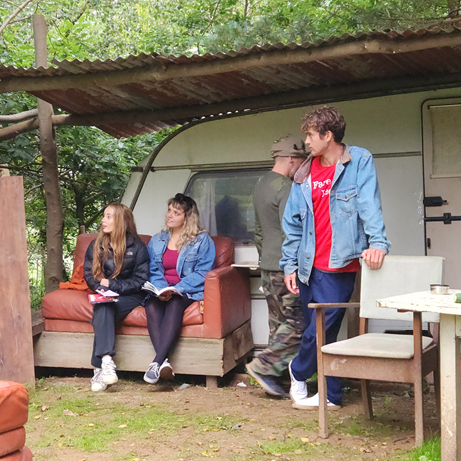 Group of people gathered outside a caravan in a wooded area, some seated and some standing, with rustic furniture around.