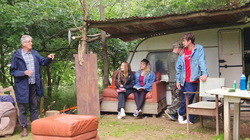 Group of people gathered outside a caravan in a wooded area, some seated and some standing, with rustic furniture around.