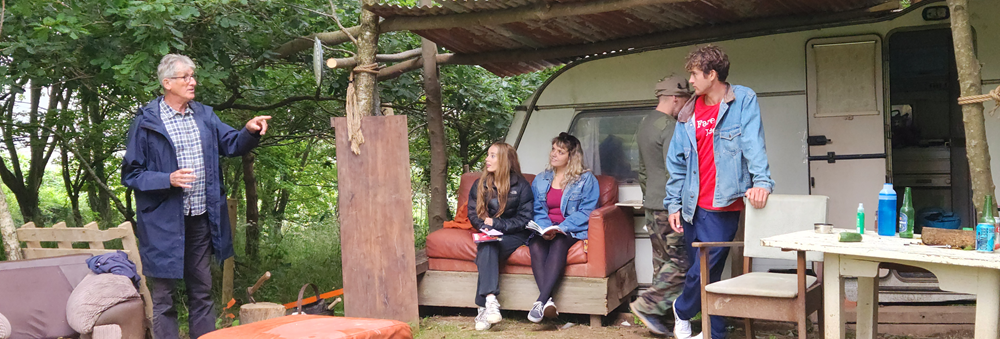 Group of people gathered outside a caravan in a wooded area, some seated and some standing, with rustic furniture around.