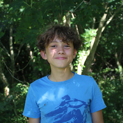 Smiling boy with curly hair wearing a blue t-shirt standing outdoors in a wooded area.