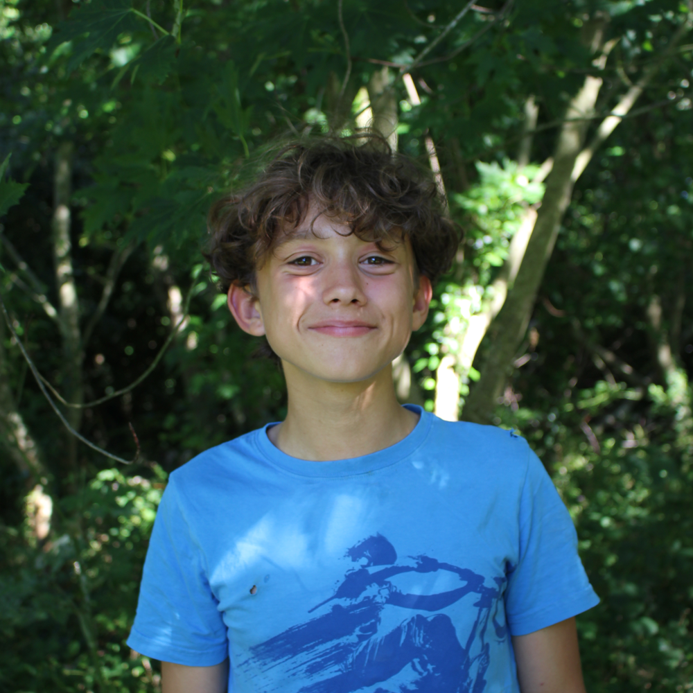 Smiling boy with curly hair wearing a blue t-shirt standing outdoors in a wooded area.