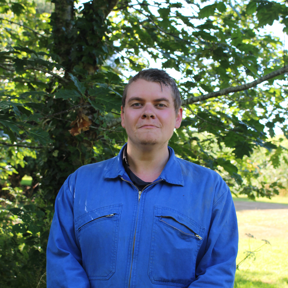 Person standing outdoors wearing a blue jacket, with green trees in the background.