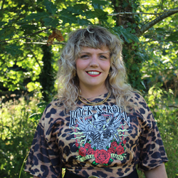 Person with curly blonde hair wearing a graphic 'Rock N' Roll' shirt with a leopard print pattern standing outdoors with greenery in the background.