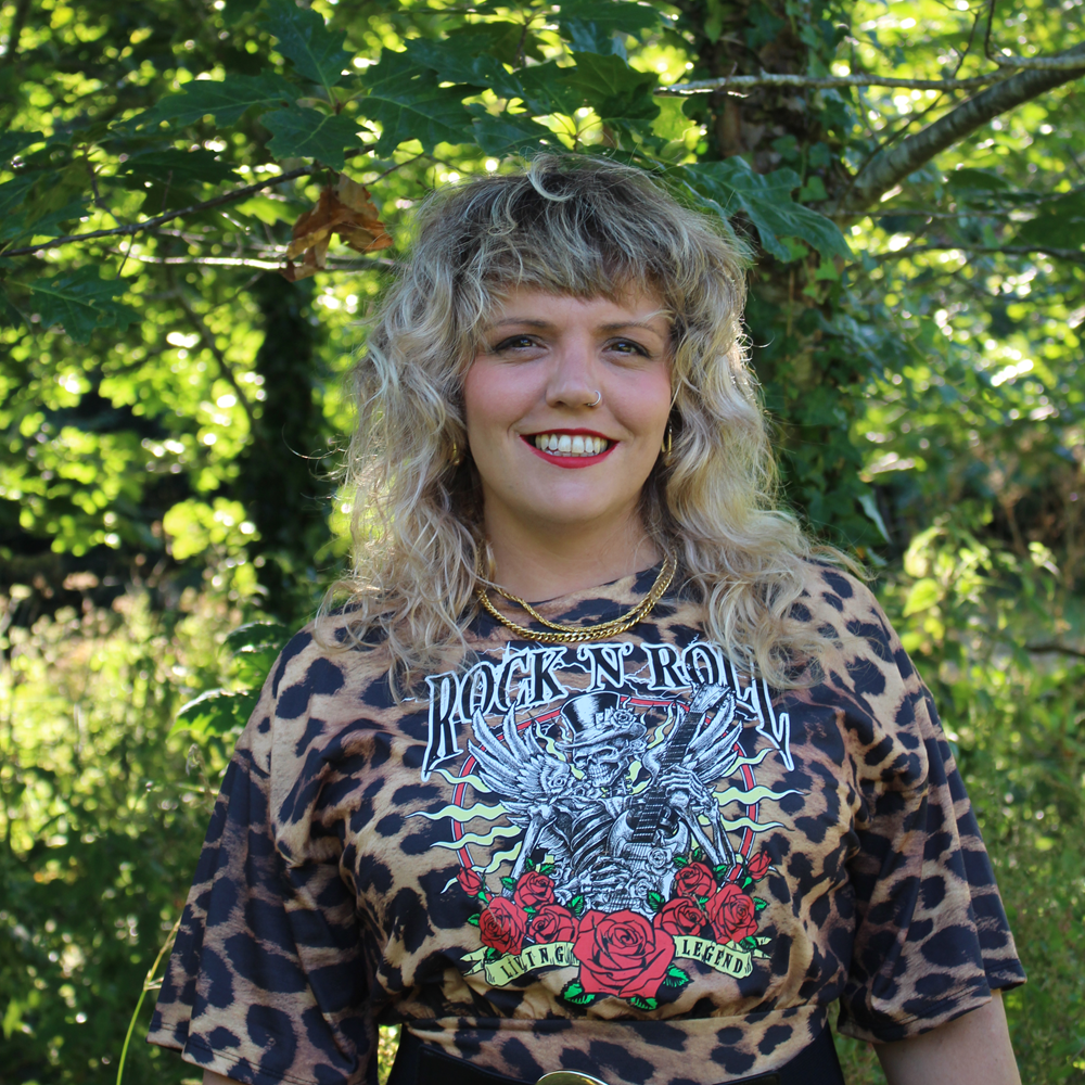 Person with curly blonde hair wearing a graphic 'Rock N' Roll' shirt with a leopard print pattern standing outdoors with greenery in the background.