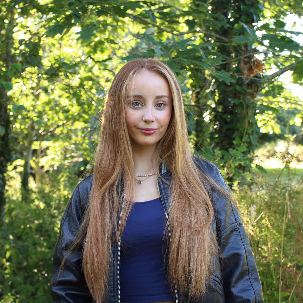 Young woman with long light brown hair standing outdoors wearing a black leather jacket