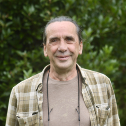Older man with long hair tied back, wearing a plaid shirt and glasses hanging around his neck, standing outside in front of green foliage.