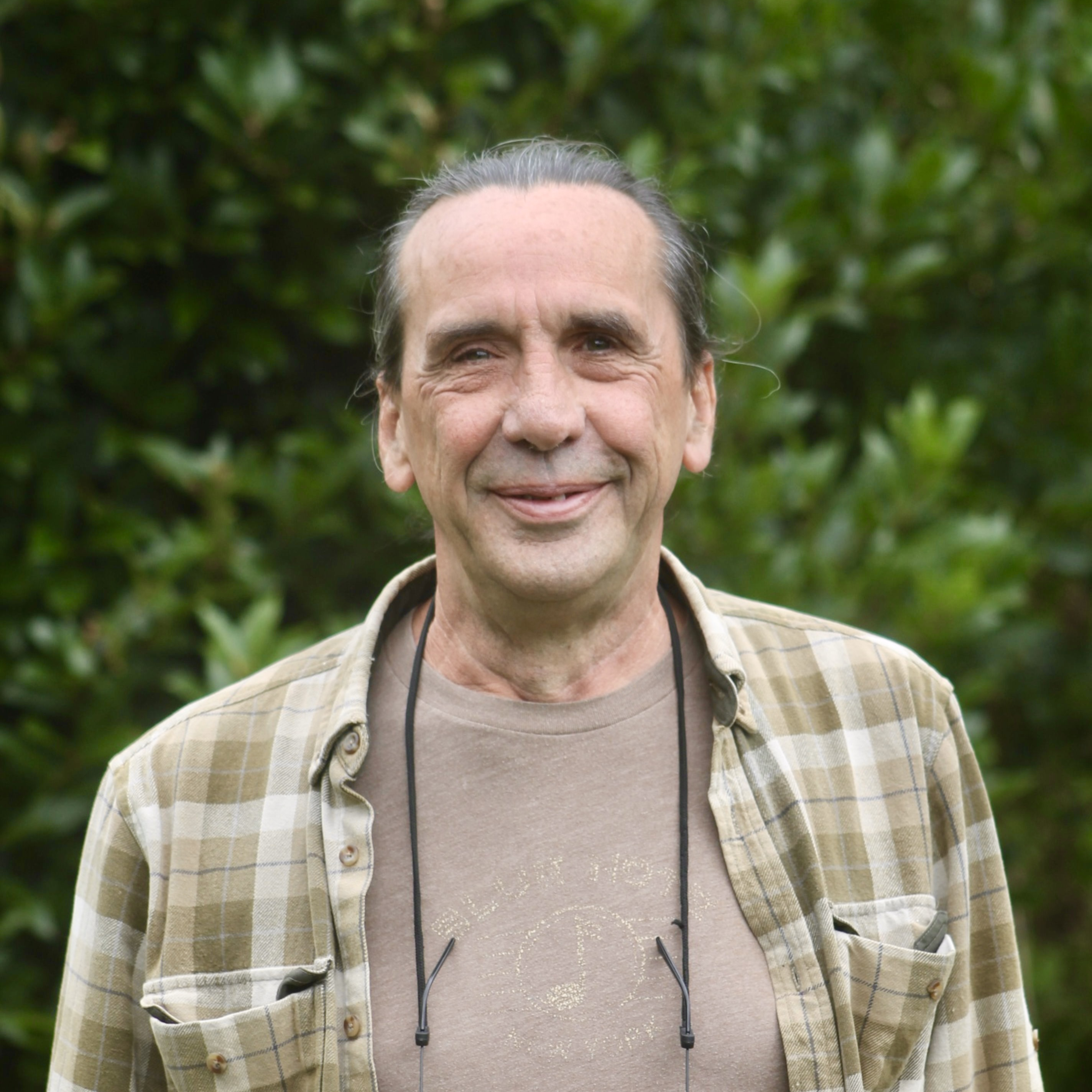 Older man with long hair tied back, wearing a plaid shirt and glasses hanging around his neck, standing outside in front of green foliage.