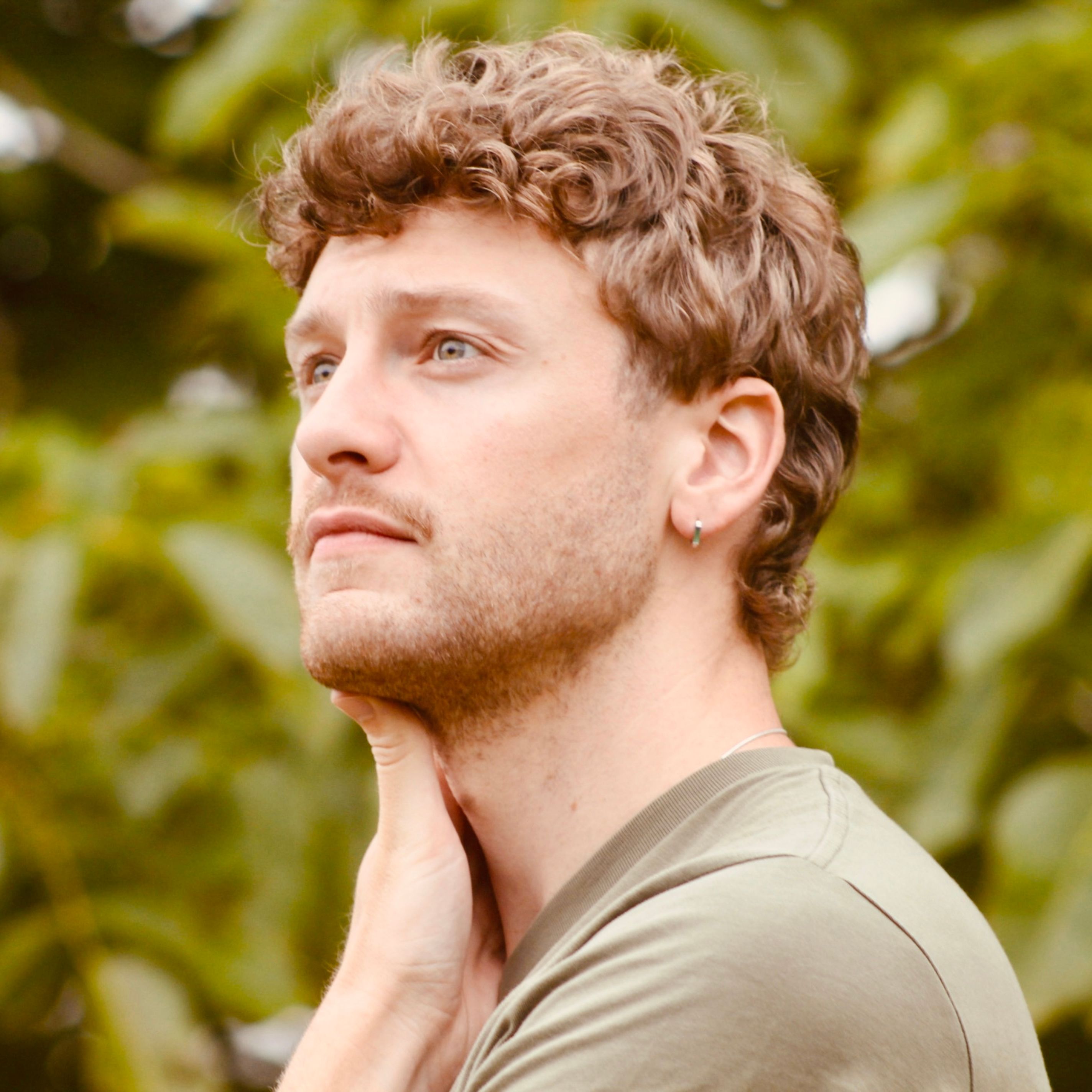 A young man with curly hair and an earring looks thoughtful outdoors, with greenery in the background.