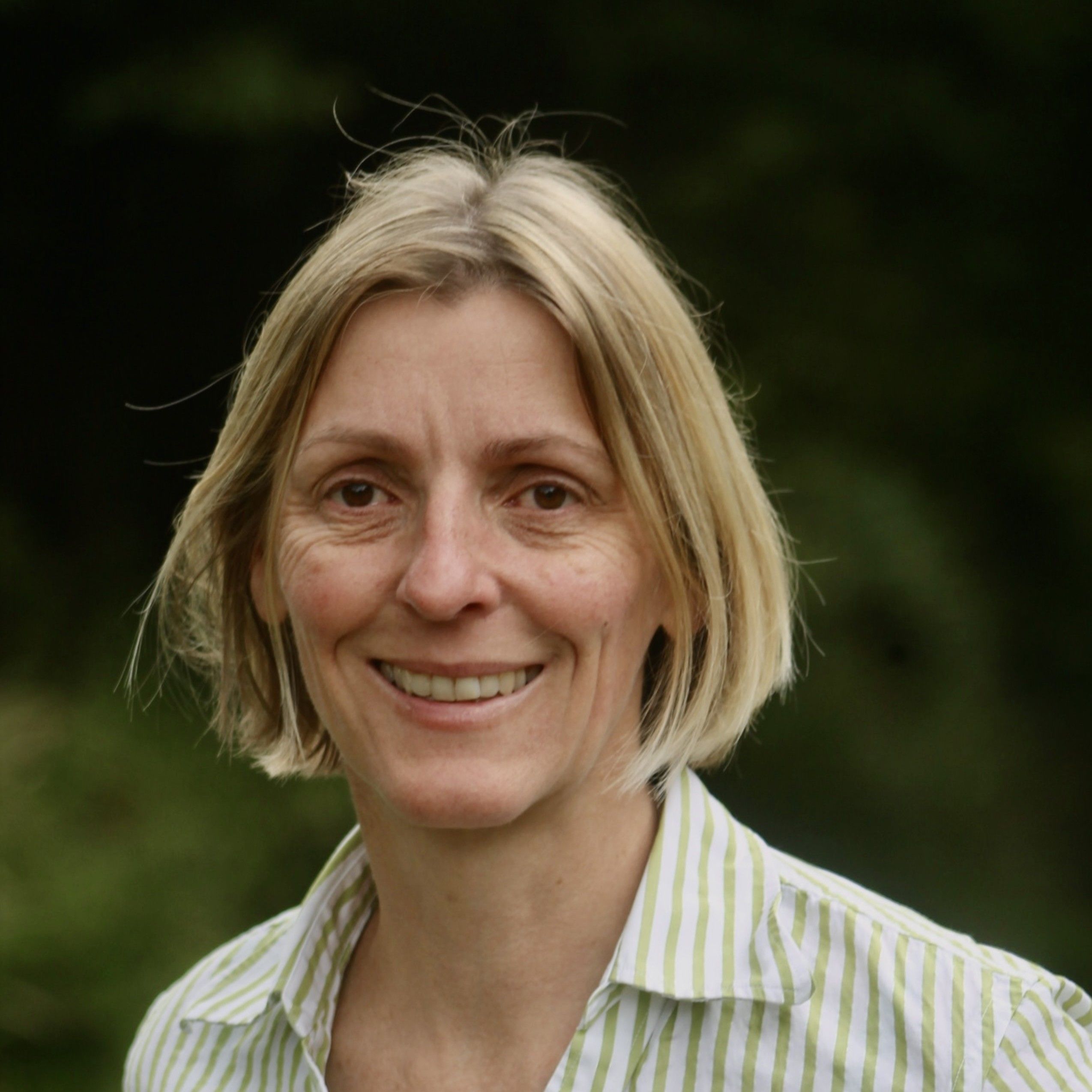 Woman with short blonde hair smiling outdoors, wearing a light-colored striped shirt