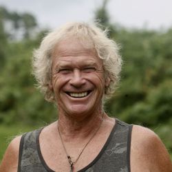 Smiling older man with curly blond hair standing outdoors, wearing a tank top and a necklace