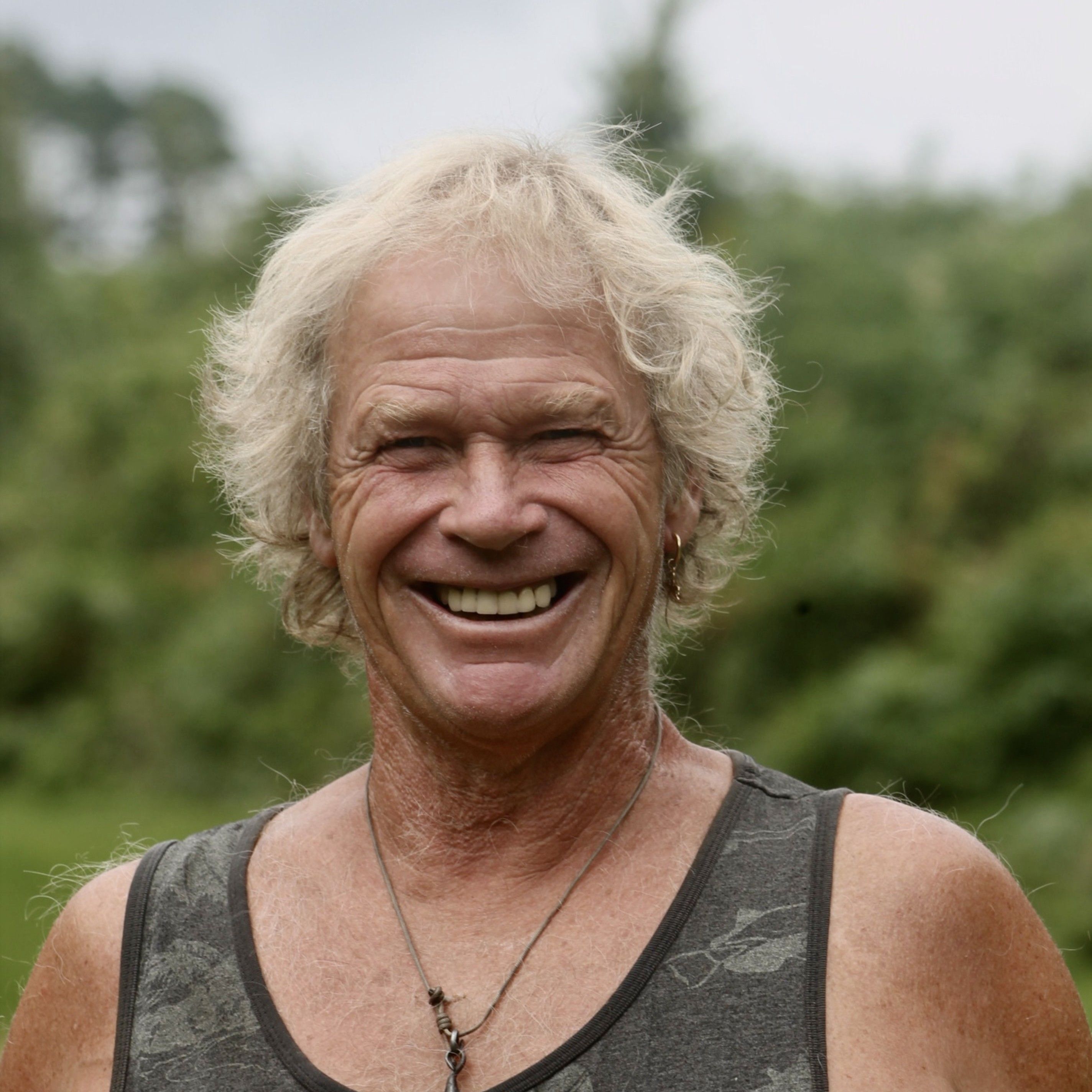 Smiling older man with curly blond hair standing outdoors, wearing a tank top and a necklace