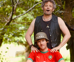 Two men outdoors, one in a leather vest with gray curly hair, the other wearing a military helmet and a red sports jersey