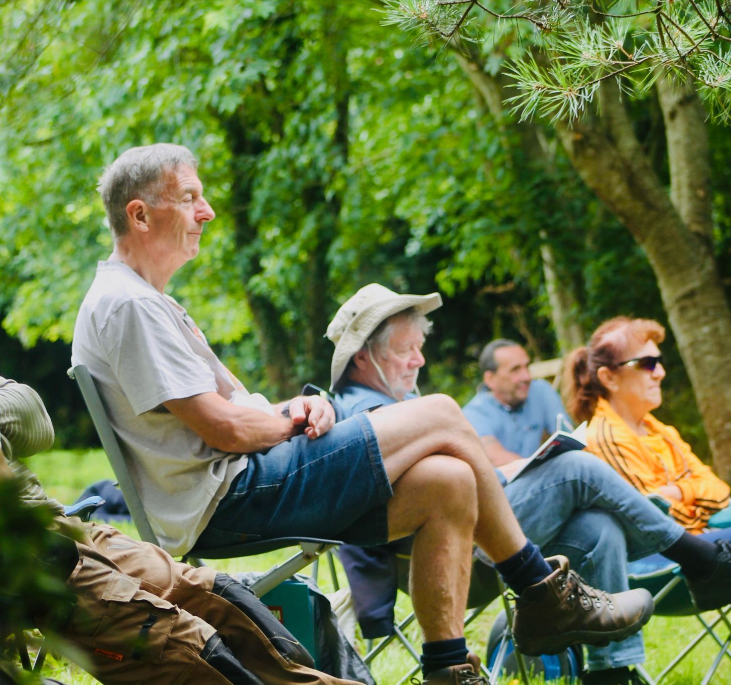 A group of older adults sitting outdoors on folding chairs, surrounded by lush green trees.