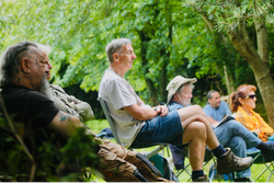 A group of older adults sitting outdoors on folding chairs, surrounded by lush green trees.