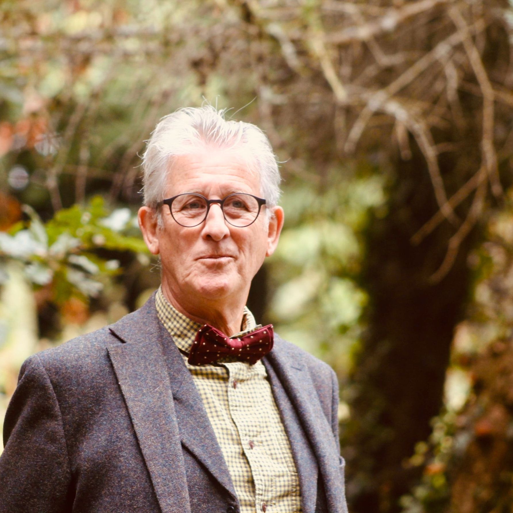 Older man in glasses and a bow tie standing outdoors among trees.
