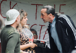 Three people in a tense conversation with a protest banner in the background.
