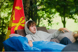 Young person in a grey hoodie sitting on an outdoor couch with a red and yellow flag and a blue tarp, surrounded by greenery.