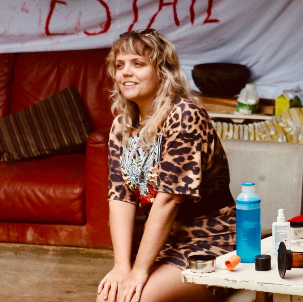Woman in a leopard print dress sitting outdoors on a chair by a cluttered table with a banner and red couch in the background.