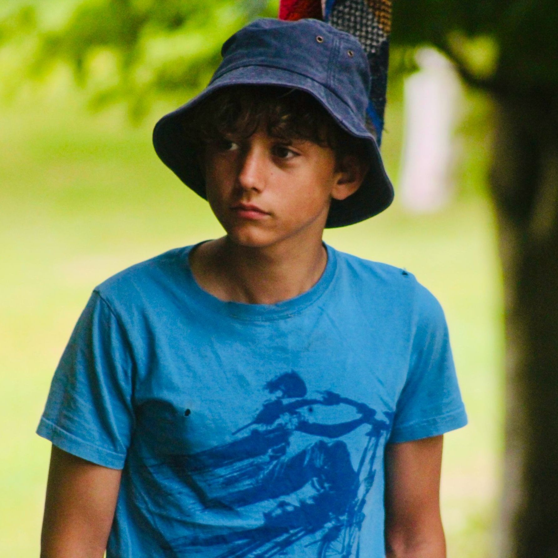 Boy wearing a blue bucket hat and blue t-shirt standing outside.