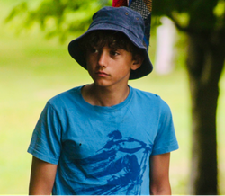 Boy wearing a blue bucket hat and blue t-shirt standing outside.